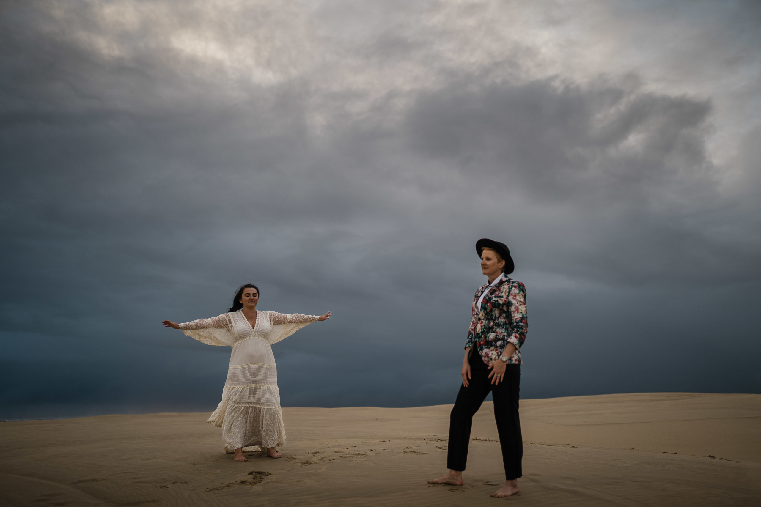 standing on top of the sand dunes during a storm, both brides dance as they are filmed for their intimate wedding film