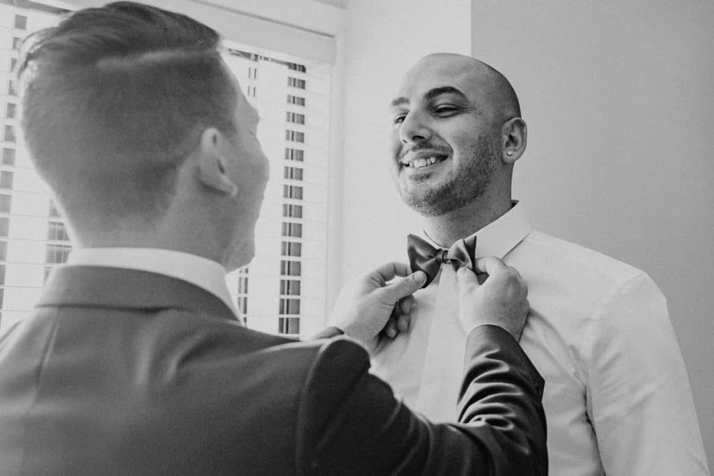 Groomsman helping Groom dress and put on bowtie before the wedding. Candid image captured by hunter valley wedding photographer james white