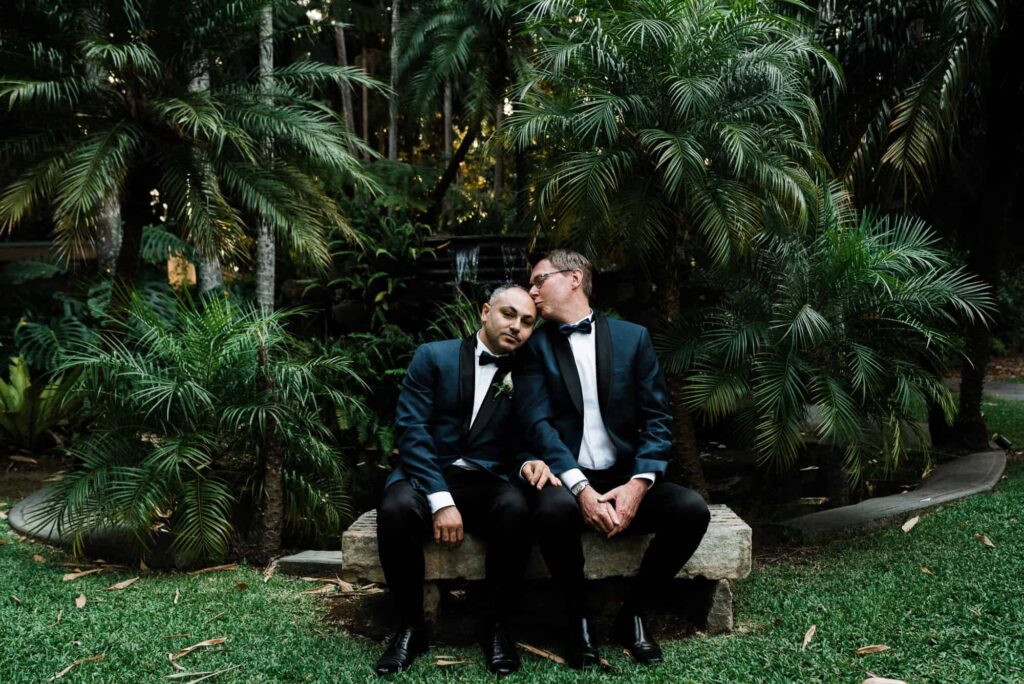 two grooms sit outside cute little chapel after wedding ceremony, surrounded by green tropical trees and a waterfall - wedding planning tips