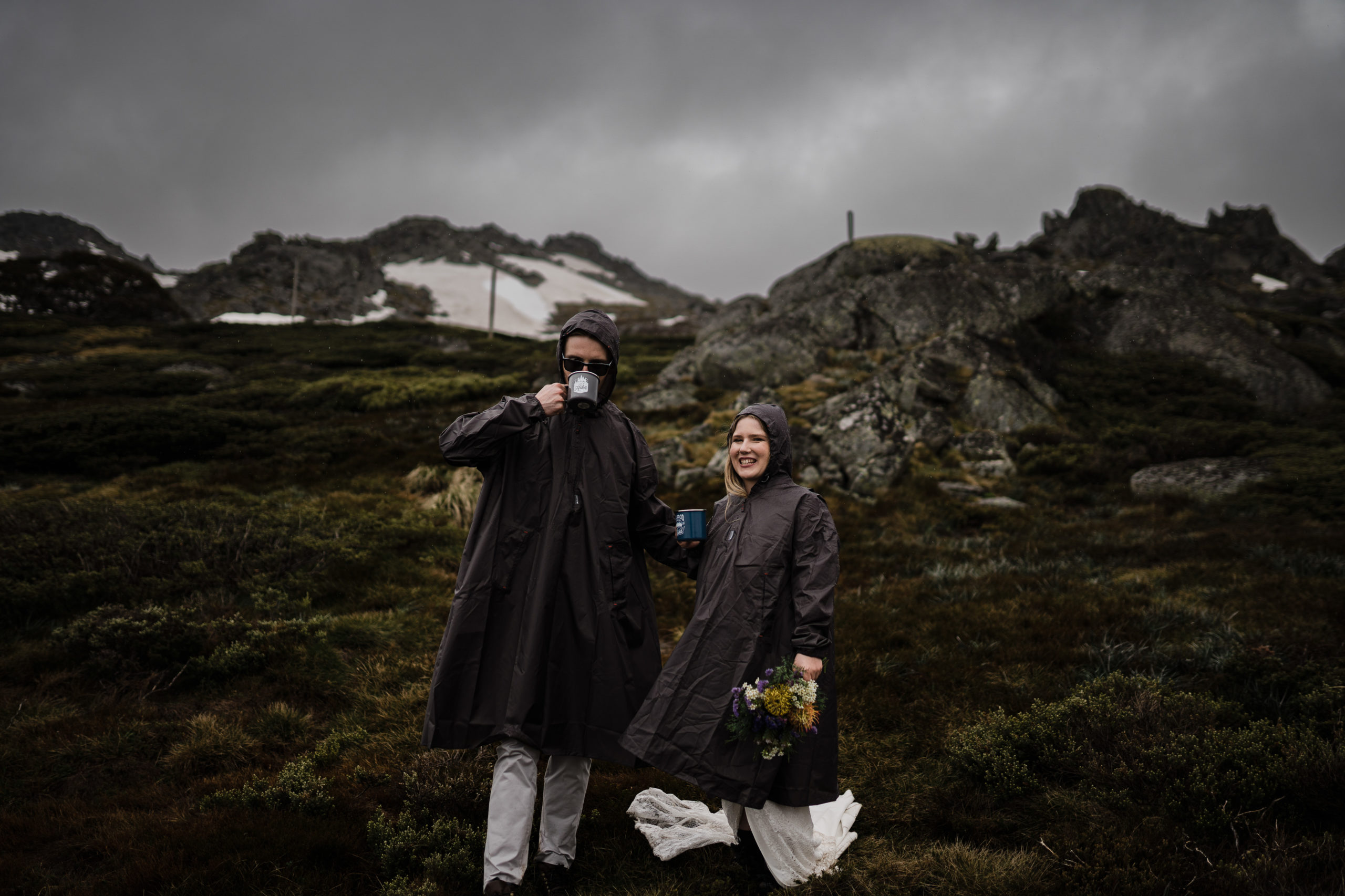bride and groom eloping on top of mount Kosciusko in the rain and snow, wearing rain coats and having a hot cup of tea during their wedding film
